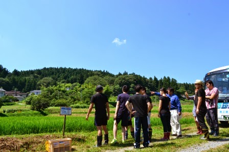 晴天の下、田んぼの前で十人ほどの人々がバスを背にして立ち、地域の風景を眺めている屋外での写真