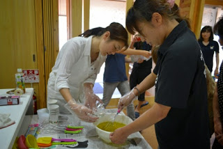 白いエプロンを着用した女性が黒いシャツを着た女性と一緒に、石けんの材料を混ぜている様子が写っている写真