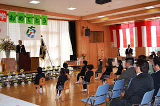 紅白幕が掛けられた会場で、演台に立つ校長らしい人物、正装した新入生たちが小さな椅子に座り、その後方に保護者たちが並んで座っている入学式の写真