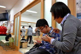 室内に設置された椅子に座った2人の親子らしき人物が、料理を箸で取りながら食事をしている様子の写真