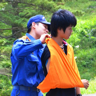 青い制服を着た消防士の男性が、黒い服を着た少年の腕をオレンジ色の三角巾で吊って応急処置の手本を見せている写真