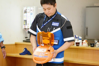 講師の男性が、オレンジ色のAED（自動体外式除細動器）の蓋を開いて持ち、受講者に使い方を説明している写真