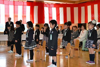 紅白幕が掛けられた会場内で、新入生の小学生が自分の席の前に立っている入学式の一コマの写真