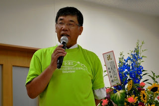 お祝いの花をバックに、緑のシャツを着た男性がマイクを手に挨拶を行っている様子をアップで撮影した写真