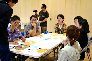 アイデアを書いた付箋やカラーペンを手にしているテーブルで、男性講師や参加者たちが笑顔で会話をしている写真