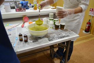 白いエプロンを着用した人が黄色い液体をお玉ですくい紙コップに注いでいて、テーブルの上にはいくつものコップと小瓶が置かれている写真