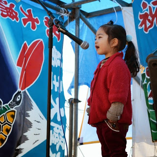 水色の大漁旗で囲まれたイベント会場のステージ上で、赤色の服を着た小さな女の子がマイクの前に立ち歌を披露している様子の写真
