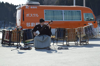 ポスクルと書かれたオレンジ色の大型ワゴン車の前で、並べて置かれた和太鼓の中央に置かれた椅子に座ったはかま姿の男性が三味線を演奏している写真