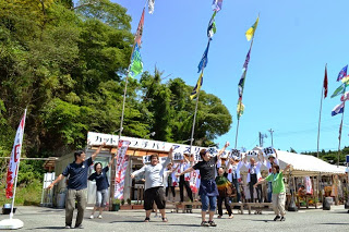 青空のもと、色とりどりの大漁旗が立つ会場でスタッフの方々が元気に踊り、後ろの人たちも音楽に合わせて踊っている写真