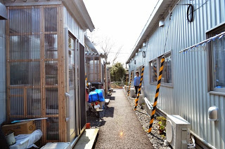 砂利敷きの細い路地を挟んだ左側に透明の波板が設置された仮設住宅の入り口に一台のバイクが停まっており、右側の住宅の入り口に女性が立っている様子の写真