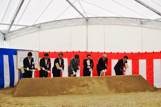 紅白幕が飾られた白いテント内で、7人のスーツ姿の男性たちが並び、山積みの砂を一斉に掘り始めている鍬入れの儀式の写真