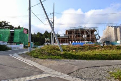 道路の奥に木造の骨組みと足場が組まれた住宅の建築現場が写っており、その隣には外壁が完成した別の建物が見える写真
