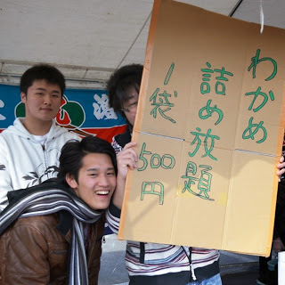 男性が「わかめ詰め放題一袋500円」と書かれた段ボール製の看板を持ち体が半分隠れるようにして持ち、他の2人の男性と一緒に写っている様子の写真