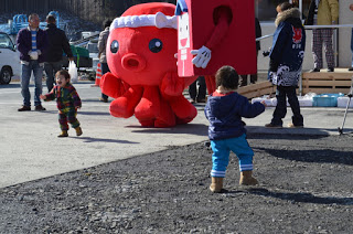 ねじり鉢巻きをしたタコのマスコットとポストのマスコットが並んで立ち、周りを小さな子どもたちが走っている様子の写真