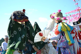 緑の布をかぶった獅子舞が踊り手の女性と向かい合っており、女性は色とりどりの着物を着て扇子を持ちながら踊っている、晴天の下で行われている祭りの様子の写真