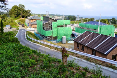 カーブした細い坂道の横に、緑色のシートで覆われた複数の建築中の住宅があり、その手前に茶色い屋根の平屋住宅がある新興住宅地を高台から撮影した写真