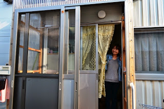 黄色のレースの暖簾が設置された仮設住宅の入り口を開け、笑顔を見せている吉田さんの写真