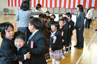 体育館の紅白幕の前で、スーツやワンピースを着た小学校の新入生たちが名札をつけて一列に並び、中にはじゃれあう児童も写っている様子の写真