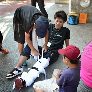 子どもと大人が協力して、白い布で別の人の左足を固定して応急処置の練習をしている写真