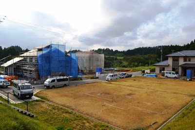 自動車が何台か停まっている道路の手前に更地が広がり、その奥で青いシートに覆われた住宅が複数建設中の写真