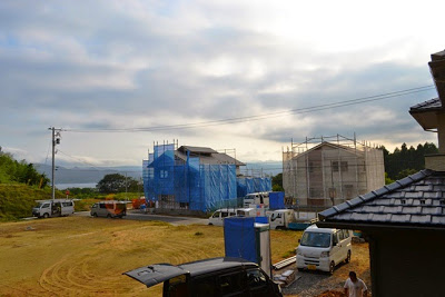 手前に自動車が数台停まり、奥に青や白のシートで覆われた住宅が海を背景に建設中である写真