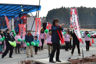 屋外イベント会場で、ピンクのシャツを着た人々が緑のポンポンを持って応援する中、赤いマフラーを首にかけた壇上の亜KIRAさんがマイクを持ち左手を前に出して歌っている様子の写真