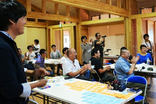 木の内装の会場で複数の人がテーブルを囲み、発表を見ながら拍手や笑顔を見せている様子の写真