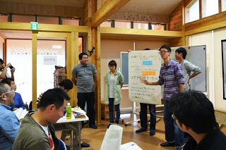 5人のグループが前に出て模造紙を掲げながら発表し、他の参加者が座って話を聞いているプレゼンテーションの写真