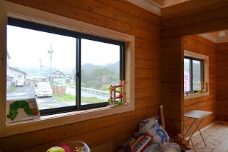 窓の外には道路と民家が見え、窓辺にはおもちゃや絵本やおもちゃが並べられている木造の室内の一角が写っている写真