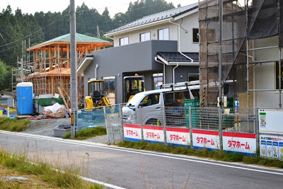 道路沿いにタマホームの看板が並ぶフェンスが設置され、手前にはトラックと重機がある複数の住宅が建設中の写真