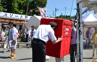 赤いポストの着ぐるみを着ようとしている人に、2人の男性が手伝って着ぐるみをかぶせている様子の写真