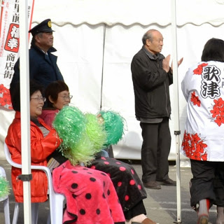 白いテントの前で行われている屋外イベントで、色違いの水玉模様のひざ掛けを足にかけている2人の女性が緑のポンポンを持って座り、立っている男性が拍手をしている様子の写真