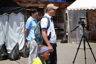 三脚に乗せたビデオカメラでイベントの様子を撮影している男性たちのそばに、青いハチマキをした男性がしゃがんでいる撮影風景の写真