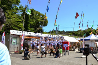 台の上に並ぶ白い法被姿のスタッフの前で、前列のスタッフの方々がポーズを決めている様子を撮影している様子の写真