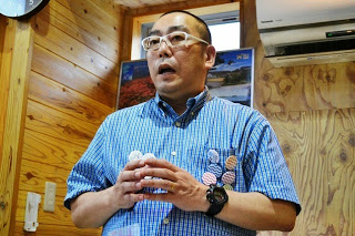 木造の室内で缶バッジを沢山つけた青色のシャツを着た男性が、真剣な表情で手を組みながら話をしている様子をアップで撮影した写真