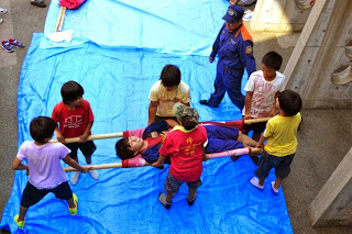 青いシートの上で男性をのせた担架を、6人の子どもたちが協力して運ぶ訓練をしている写真