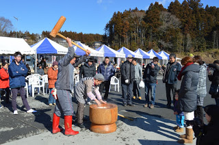 白と青の屋根のテントが立ち並ぶ前で、木製の杵と臼で餅つきが行われ、その周りを大勢の来場者が囲んで見物している写真
