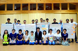 背景の木の壁の前で、模造紙を持った笑顔の参加者が横2列に並んでいる集合写真