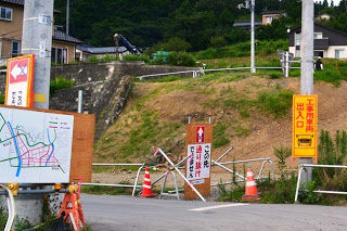 地図の看板と「工事用車両出入口」「この先通り抜けできません」などの警告標識が設置され、背後に草が生い茂る斜面と住宅が見える写真