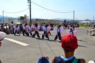 日傘をさした観客が見ているなか、お揃いの白い法被を着た子どもたちが2列に並び、両手と両足を大きく広げてダンスを行っている、よさこいステージの様子の写真