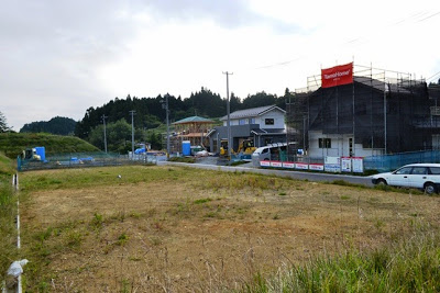 白い自動車が停まっている道路の両側の更地の奥に、足場とシートで覆われた建設中の複数の住宅が並んでいる写真