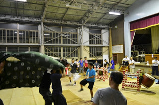 体育館の中で太鼓や笛を演奏する人たちと、獅子舞の練習をしている人々が集まっている様子が写っている写真