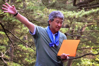 グレーのシャツを着た男性がオレンジ色のファイルを持ちながら右手を上げて話している屋外での説明の写真