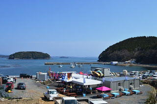 海辺に屋台が立ち並び、その近くに車が停められていて、左奥には青島が見え、海には小島が見える風景を写した写真。