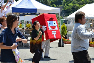 赤いポスト型のキャラクター「ポストくん」が周囲の人たちと一緒に楽しそうにダンスしている様子の写真