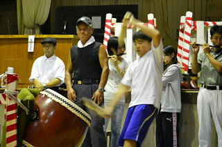 大人たちが見守りながら、後ろで演奏している笛の音色に合わせて、全力で和太鼓を叩いている男の子の写真