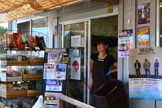 周囲にお菓子やポスターが並んでいる商店街の店舗の入口で、エプロン姿の女性が立ちダンスを見ている写真