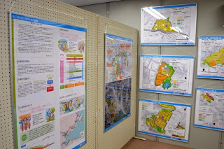 有孔ボードや壁に掲示された複数の地図や情報ポスターが写っており、左側には図や文章が記載された大きなポスター、右側には色分けされた地域を示す地図が並ぶ展示資料の写真