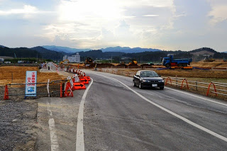 道路に黒い車が走っており、奥には土を盛った工事現場と重機が見え、道路脇にオレンジ色の矢印標識が並んでいる道路工事中の写真