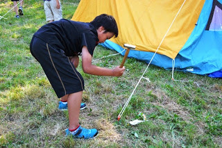 黒い服を着た少年がハンマーでテントのロープを地面に打ちつけて設営作業をしている写真